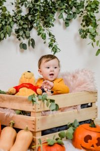 Cute baby sitting in a wooden crate with pumpkins and plush toy, decorated for Halloween.