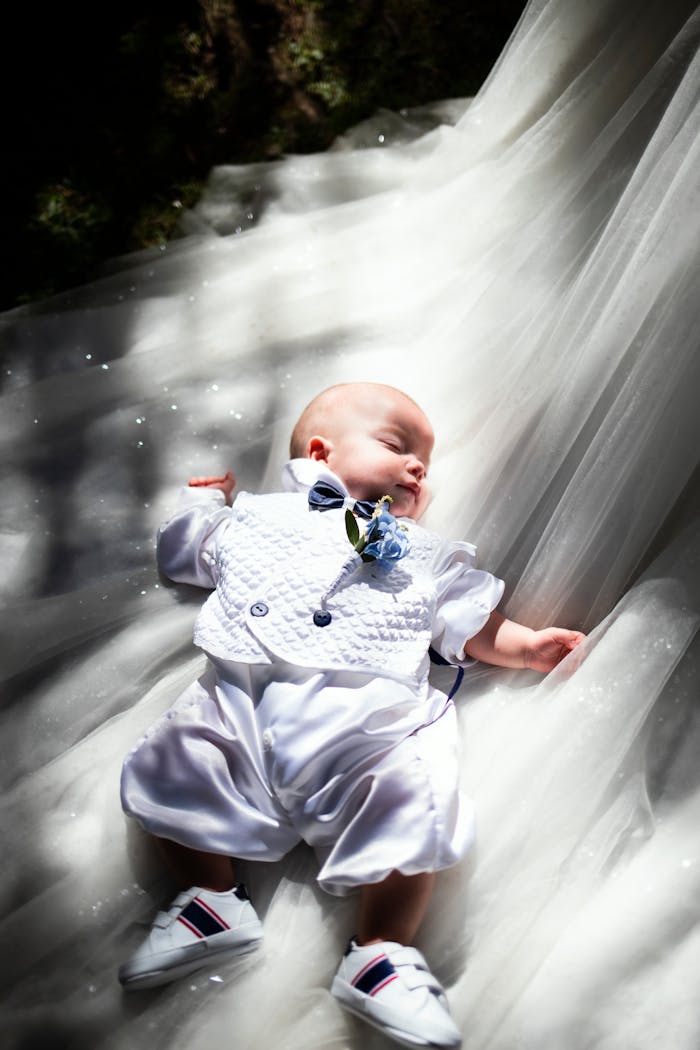 services-04 Charming baby dressed in elegant white clothes lying peacefully on tulle fabric in natural light.