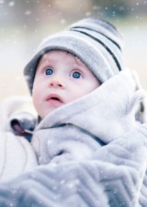 Cute baby wrapped in a blanket with a knit hat, gazing upward during snowfall.